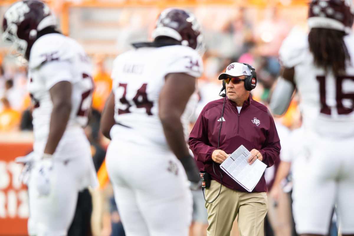 Jimbo Fisher walks on the field holding papers and wearing a headset